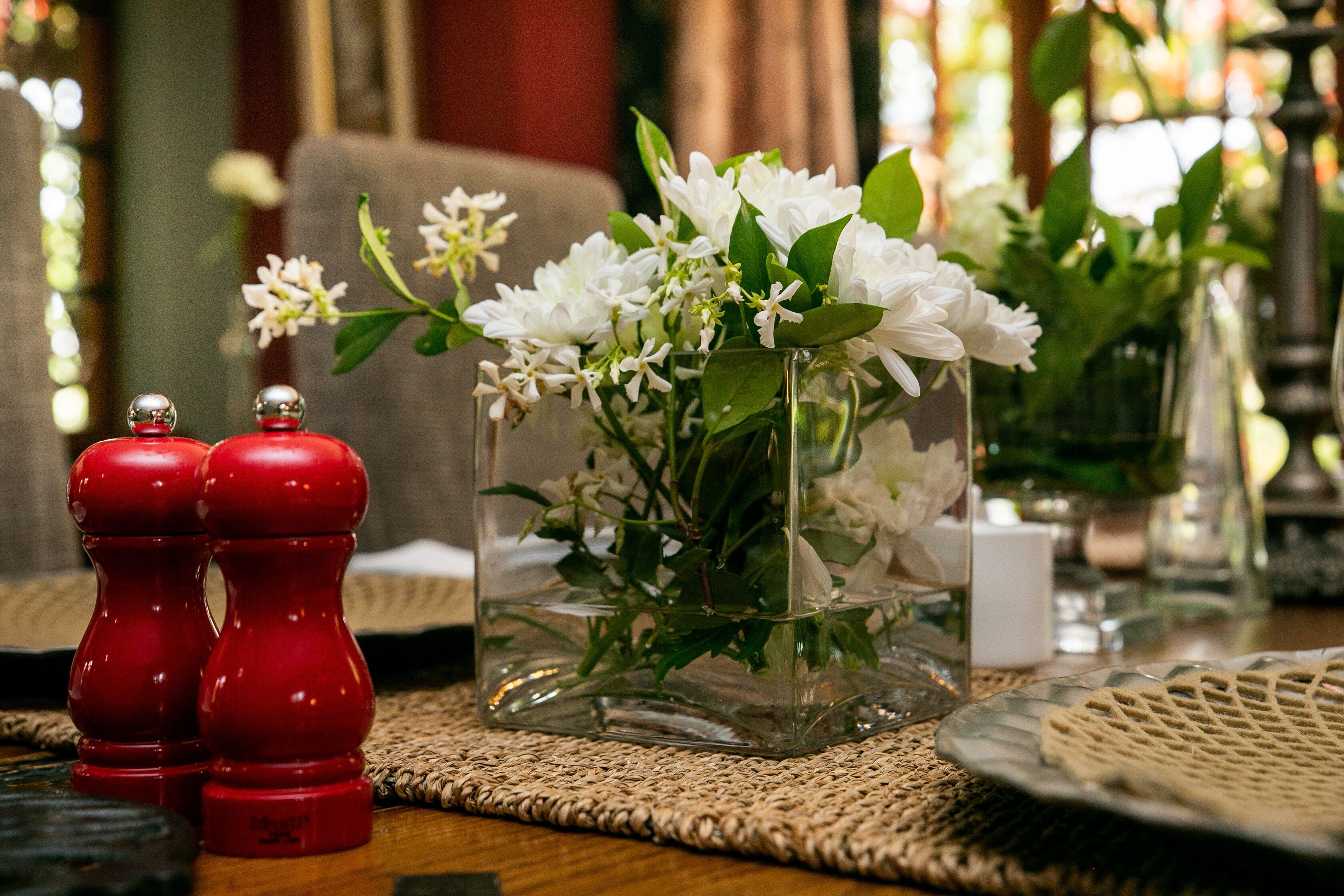 A table with a clear glass vase containing white flowers and green leaves, a woven placemat, and two red salt and pepper shakers.