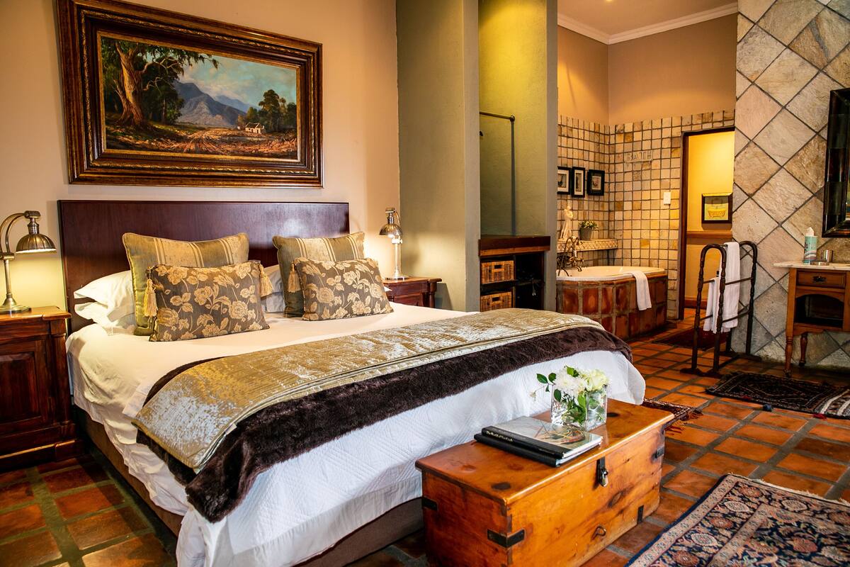 A hotel room with a large bed, a wooden headboard, a nightstand, a bedside table, a mirror, a bathroom with a sink and a towel rack, and a framed landscape painting on the wall.