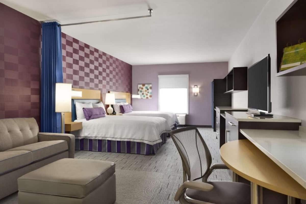 A hotel room with a checkered wall, a bed, a desk, a chair, a sofa, and a TV.