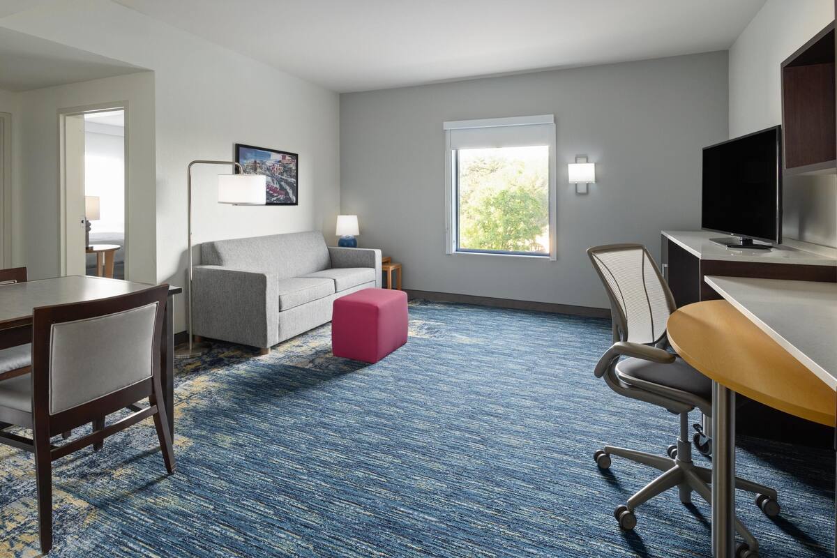 A hotel room with a grey sofa, a pink ottoman, a dining table with chairs, a TV, and a large window with a view of greenery.