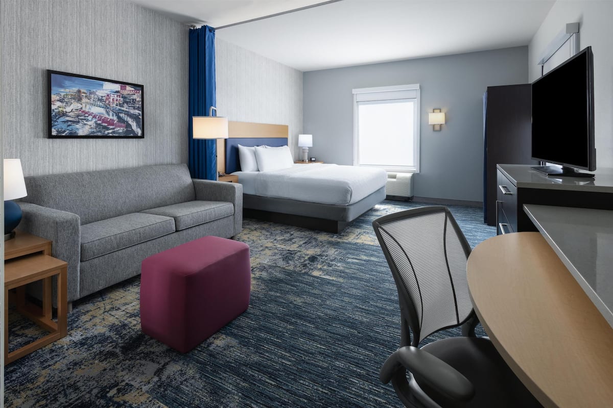 A hotel room with a bed, a sofa, a desk, a TV, and a painting on the wall.