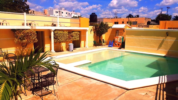 Una piscina con zona de terraza, rodeada de edificios con paredes amarillas y un cielo azul despejado.
