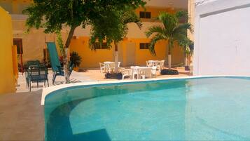 Un área de piscina con una piscina azul cristalina, rodeada de sillas y mesas de plástico blanco y palmeras.