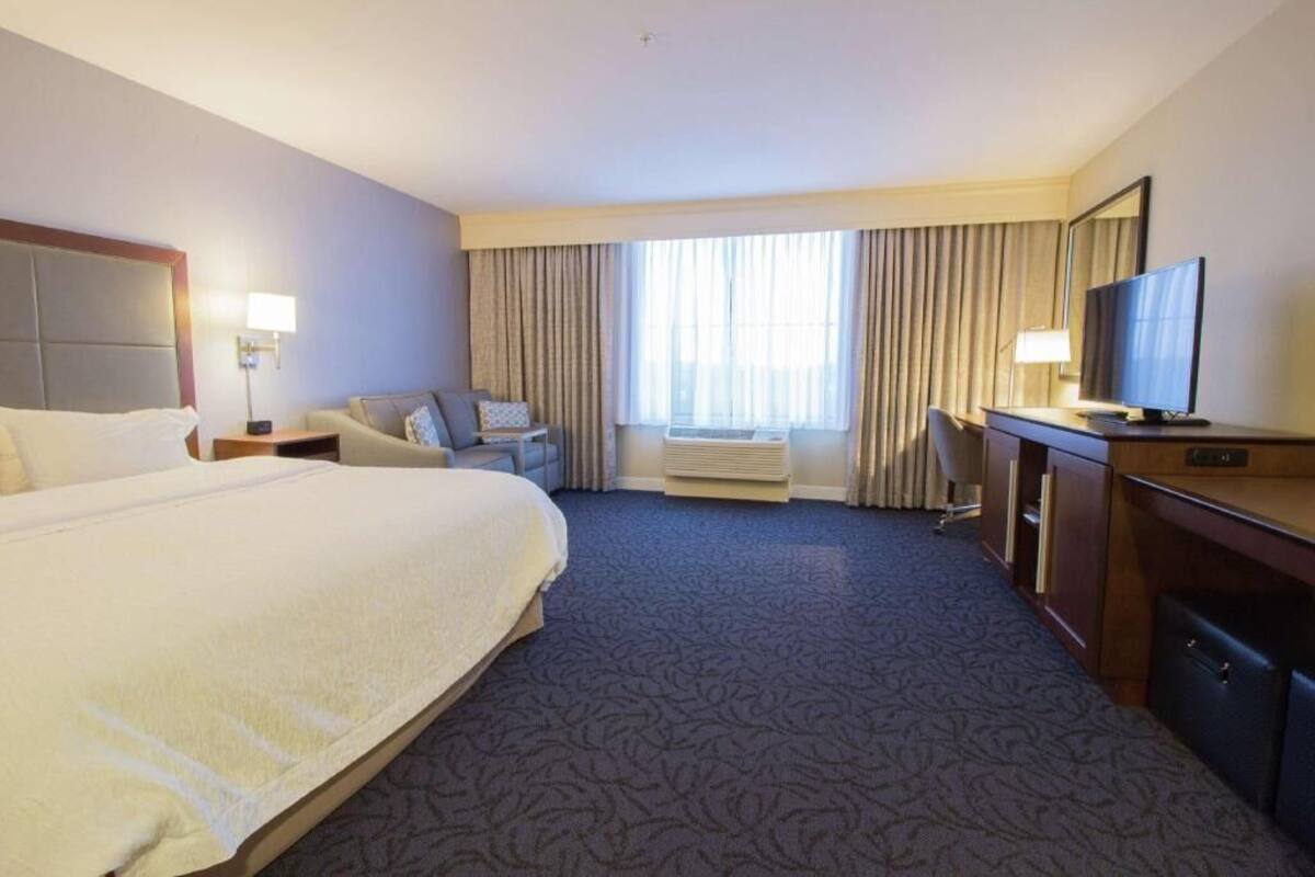 A hotel room with a large bed, a desk with a television, a chair, a desk lamp, and a window with curtains.