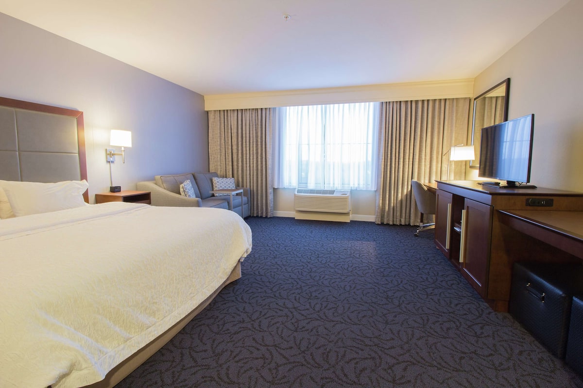 Une chambre d’hôtel avec un grand lit, un bureau avec une télévision, une chaise, une lampe de bureau et une fenêtre avec des rideaux.