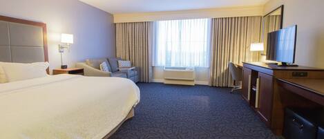 A hotel room with a large bed, a desk with a television, a chair, a desk lamp, and a window with curtains.