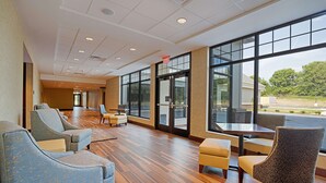A spacious lobby with a wooden floor, comfortable seating, large windows, and a glass door leading outside.