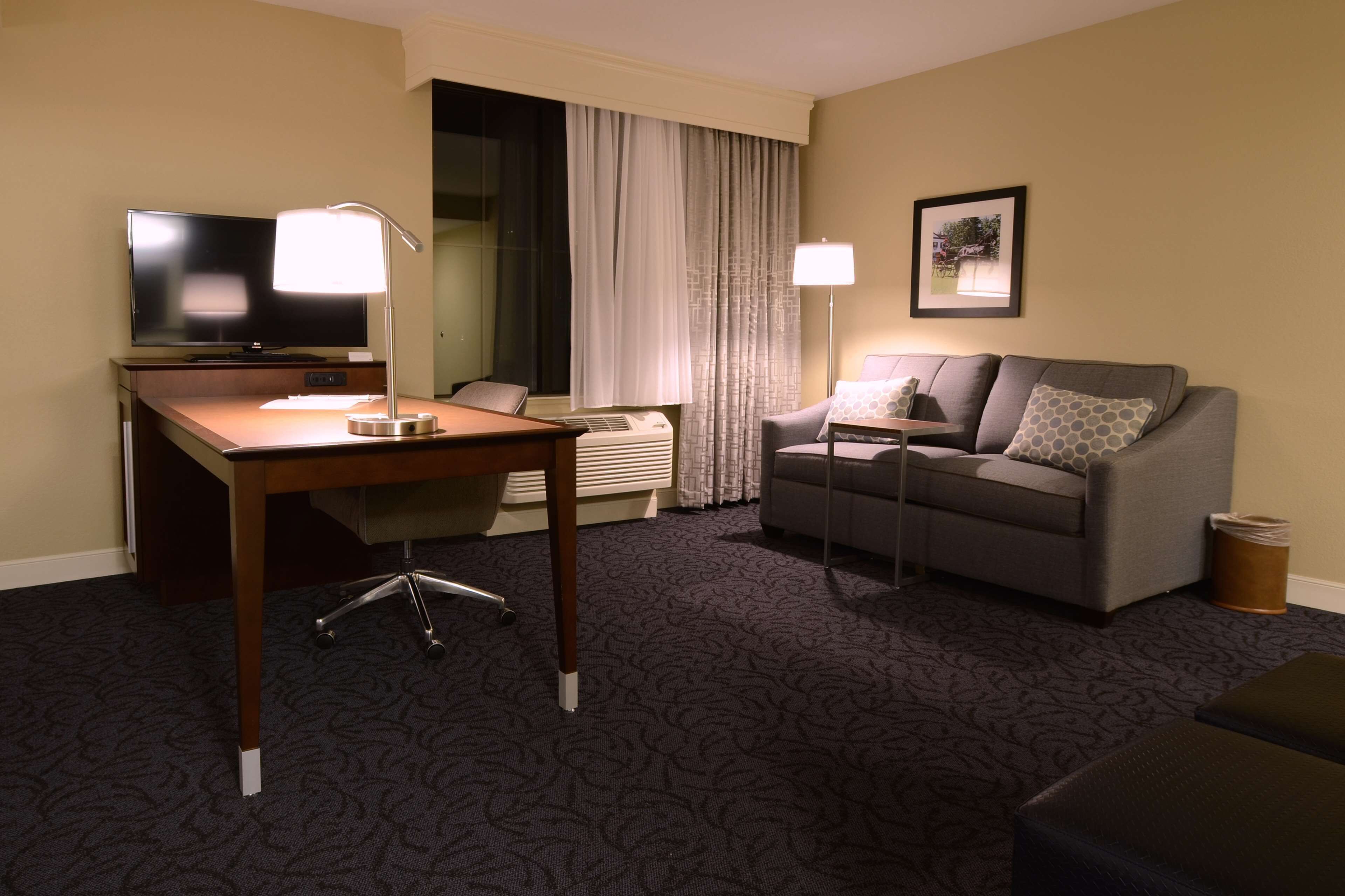 A hotel room with a wooden desk, a television, a grey sofa, and a patterned carpet.