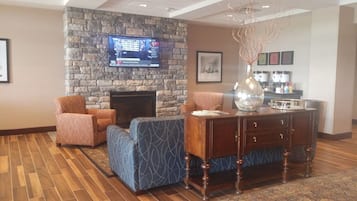 A lobby with a stone fireplace, two patterned armchairs, a blue sofa, a wooden coffee table, and a flat-screen TV displaying sports content.