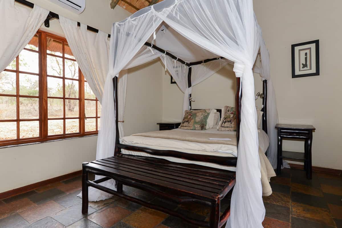 Una cama con dosel y cuatro columnas, un banco en frente, una mesita de noche y una ventana con cortinas.