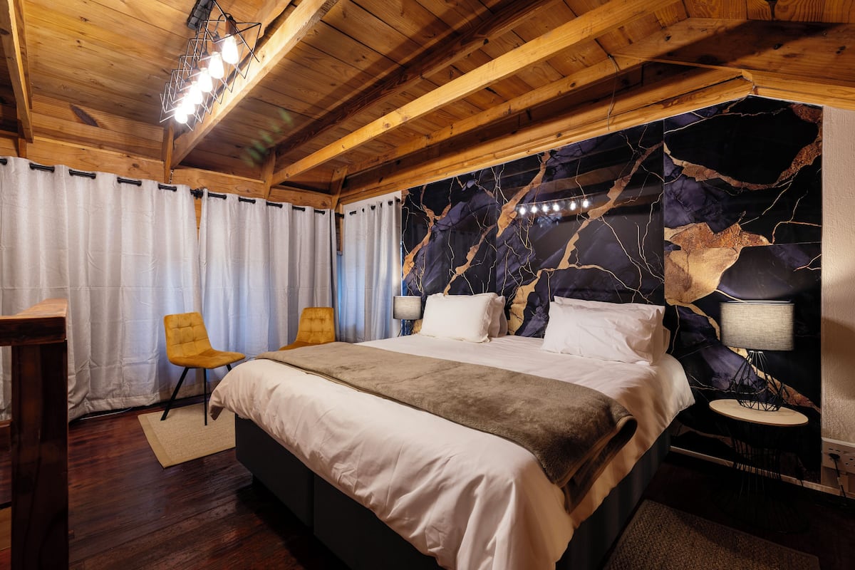 Techo de madera, cama con ropa de cama blanca, una silla amarilla, mesita de noche con lámpara y pared con diseño tipo mármol.