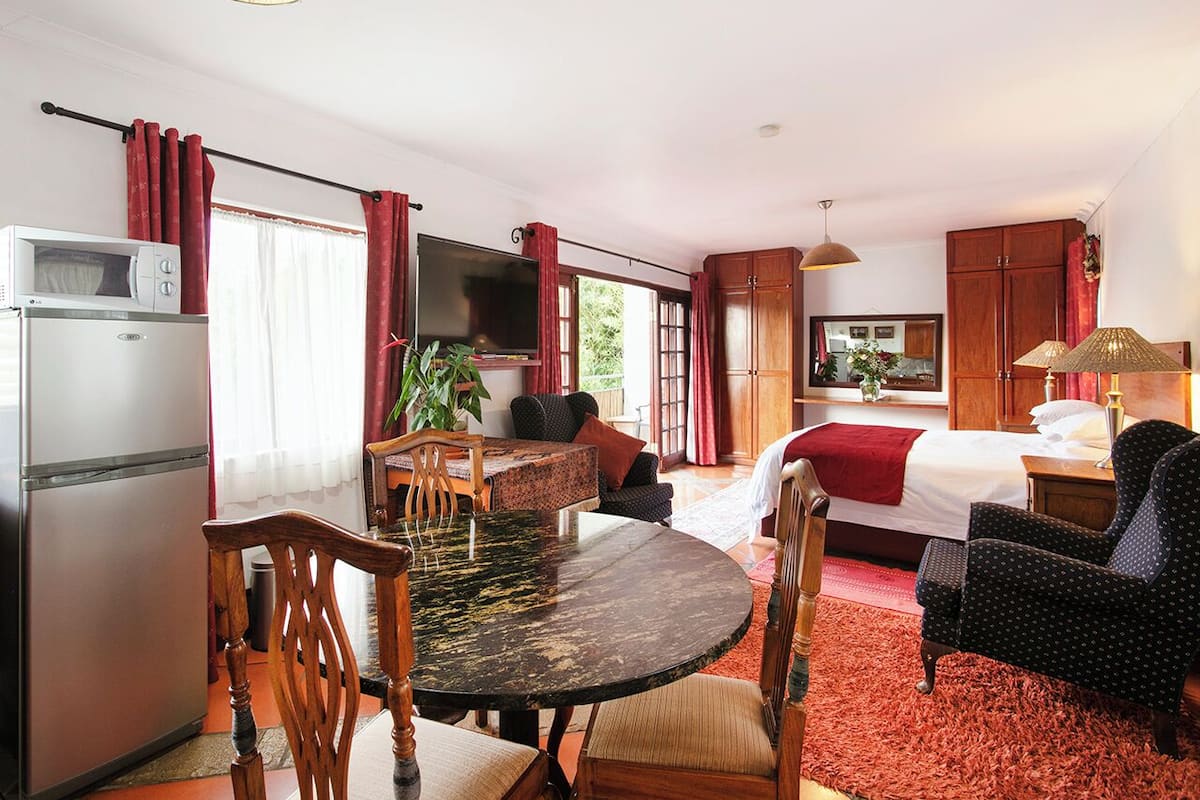 Una habitación con cama, televisión, mesa de comedor con sillas, zona de cocina con refrigerador y ventana con cortinas.