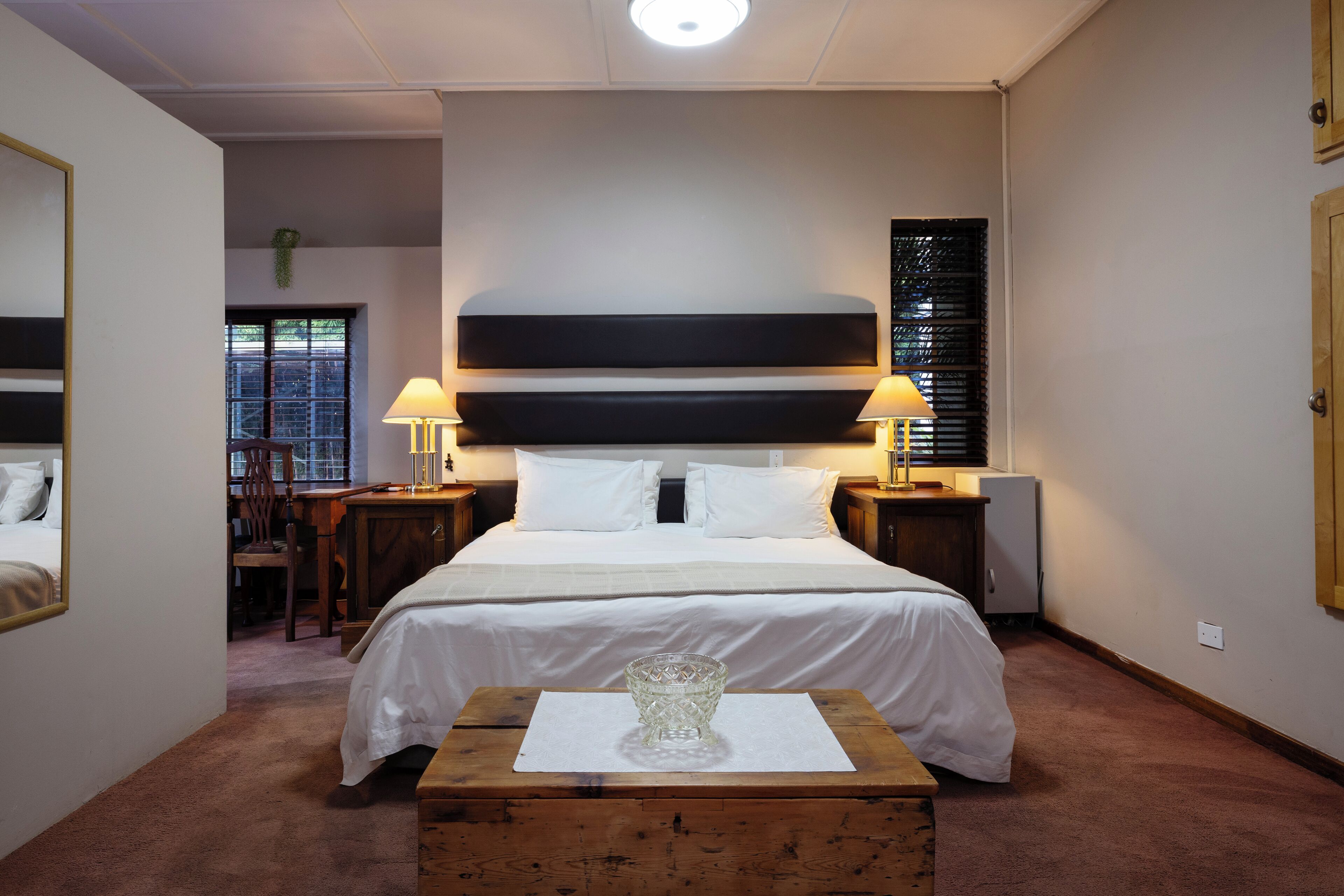 A neatly arranged bedroom with a bed, bedside lamps, a small table with a glass, and a window with blinds.