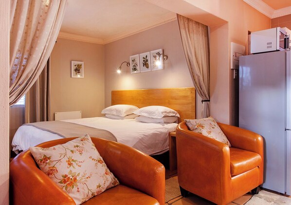 A hotel room with a bed, two armchairs, a nightstand, a wall-mounted lamp, a refrigerator, and a wall with three framed pictures.