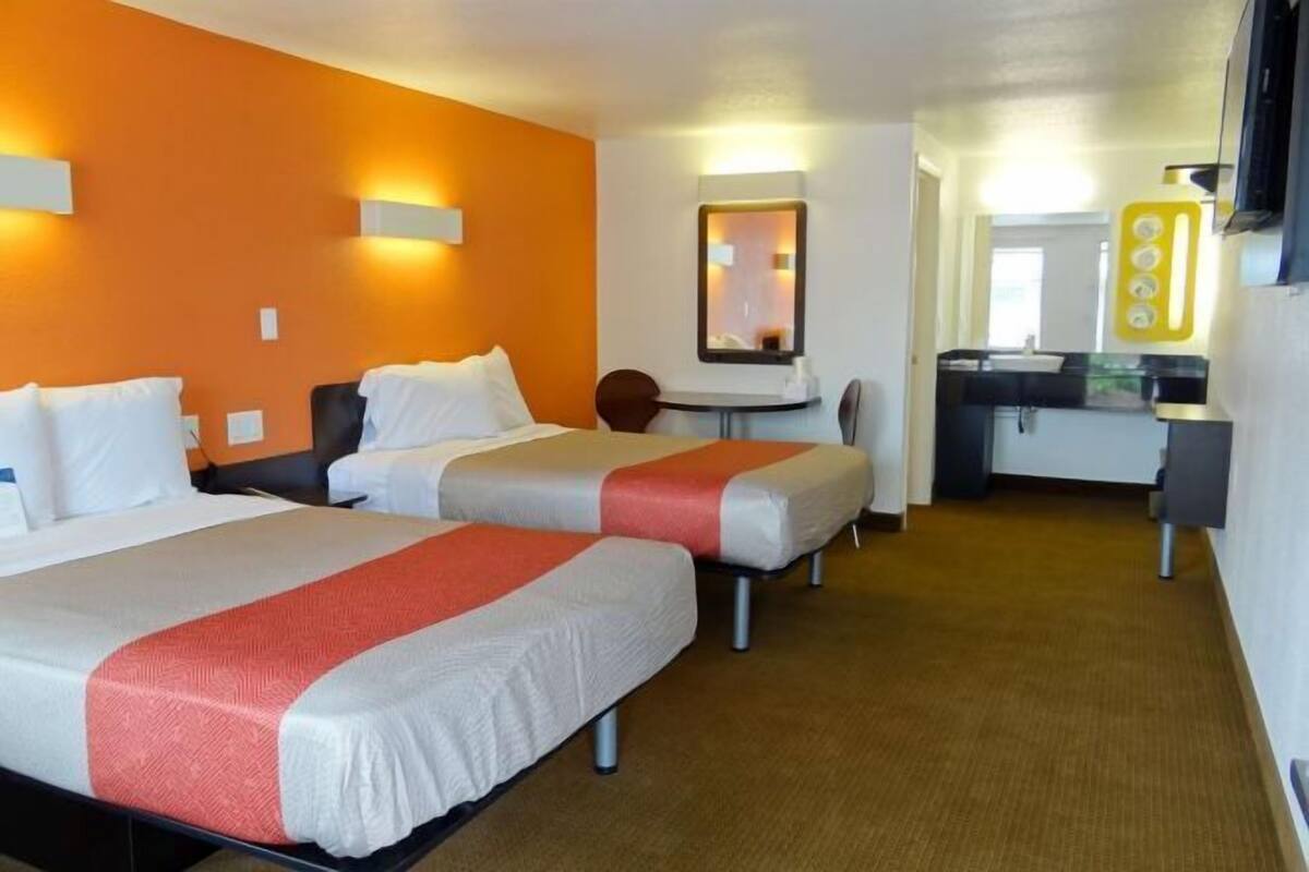 A hotel room with two beds, a small desk, a mirror, and a kitchenette.
