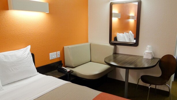A hotel room with a bed, a small table, a chair, a mirror, and a wall-mounted light. - Motel 6 Erie, PA (Erie)