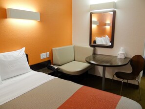 A hotel room with a bed, a small table, a chair, a mirror, and a wall-mounted light.