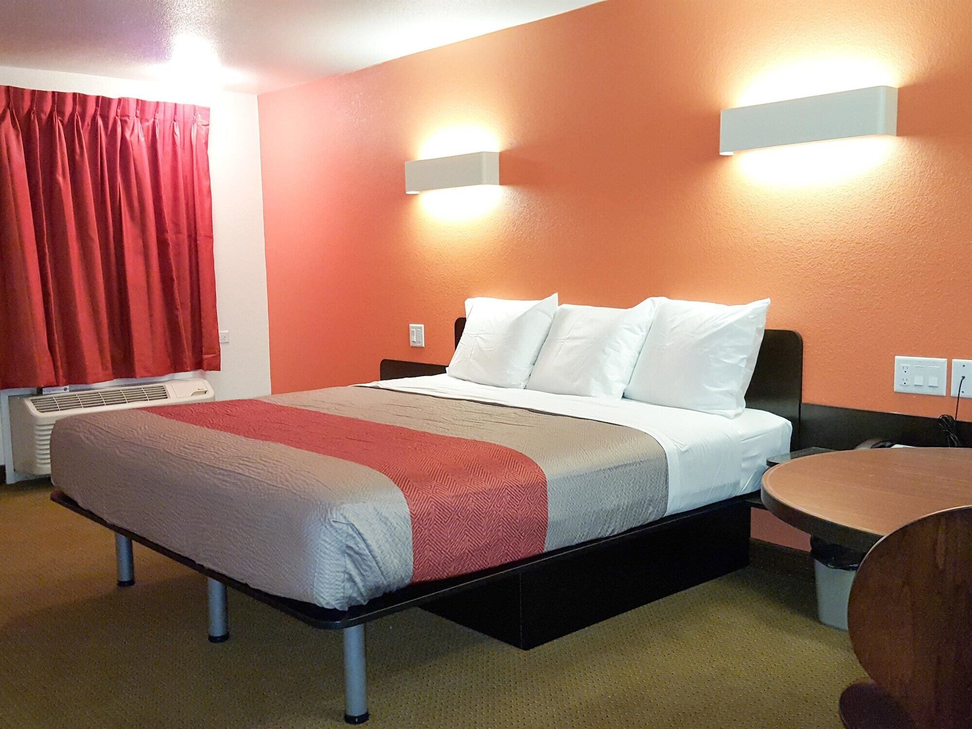 A hotel room with a bed, a bedside table, a wall-mounted light, and a window with red curtains.
