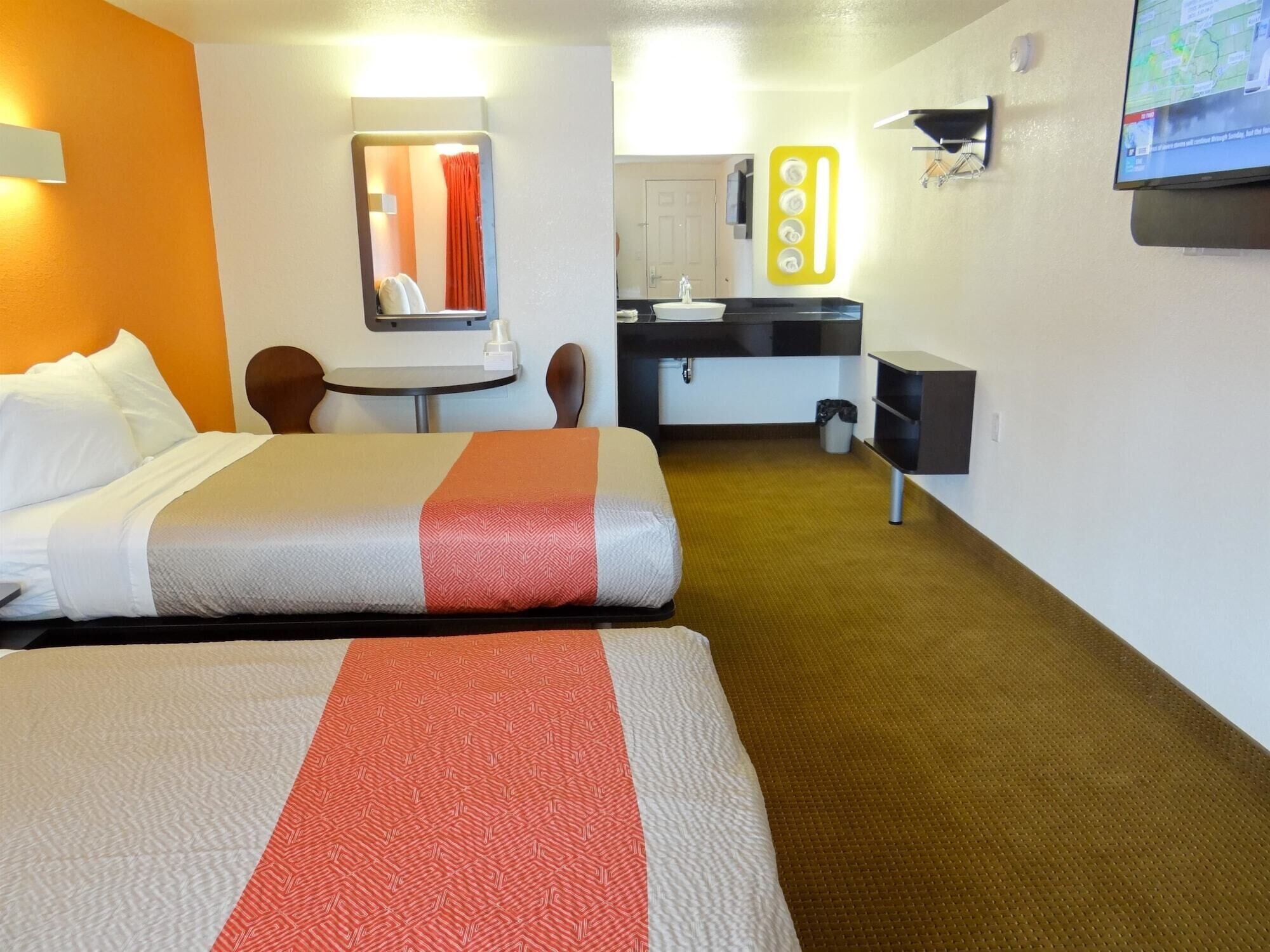 A hotel room with two beds, a small table, a chair, a sink, and a television.