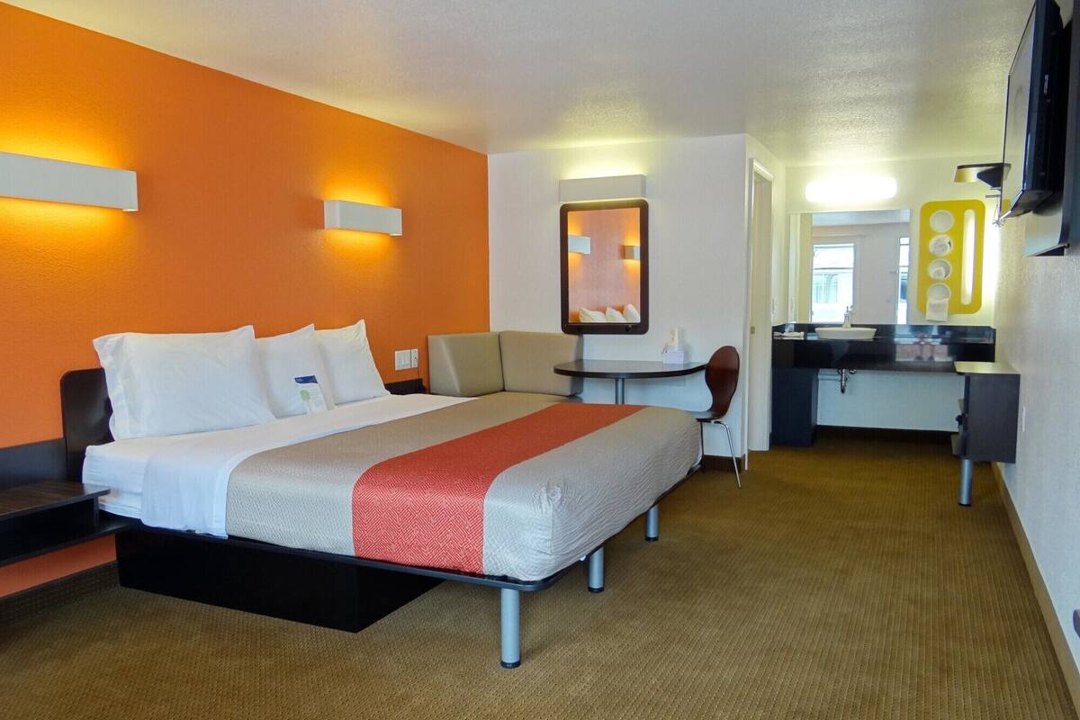 A hotel room with a bed, a desk, a chair, a small table, a mirror, and a bathroom with a sink and mirror.