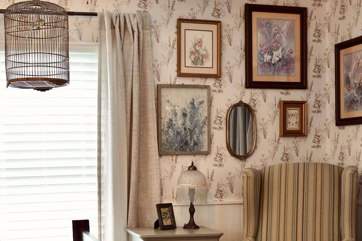 Quarto com cama de madeira, poltrona listrada, mesa de cabeceira com abajur e papel de parede floral.