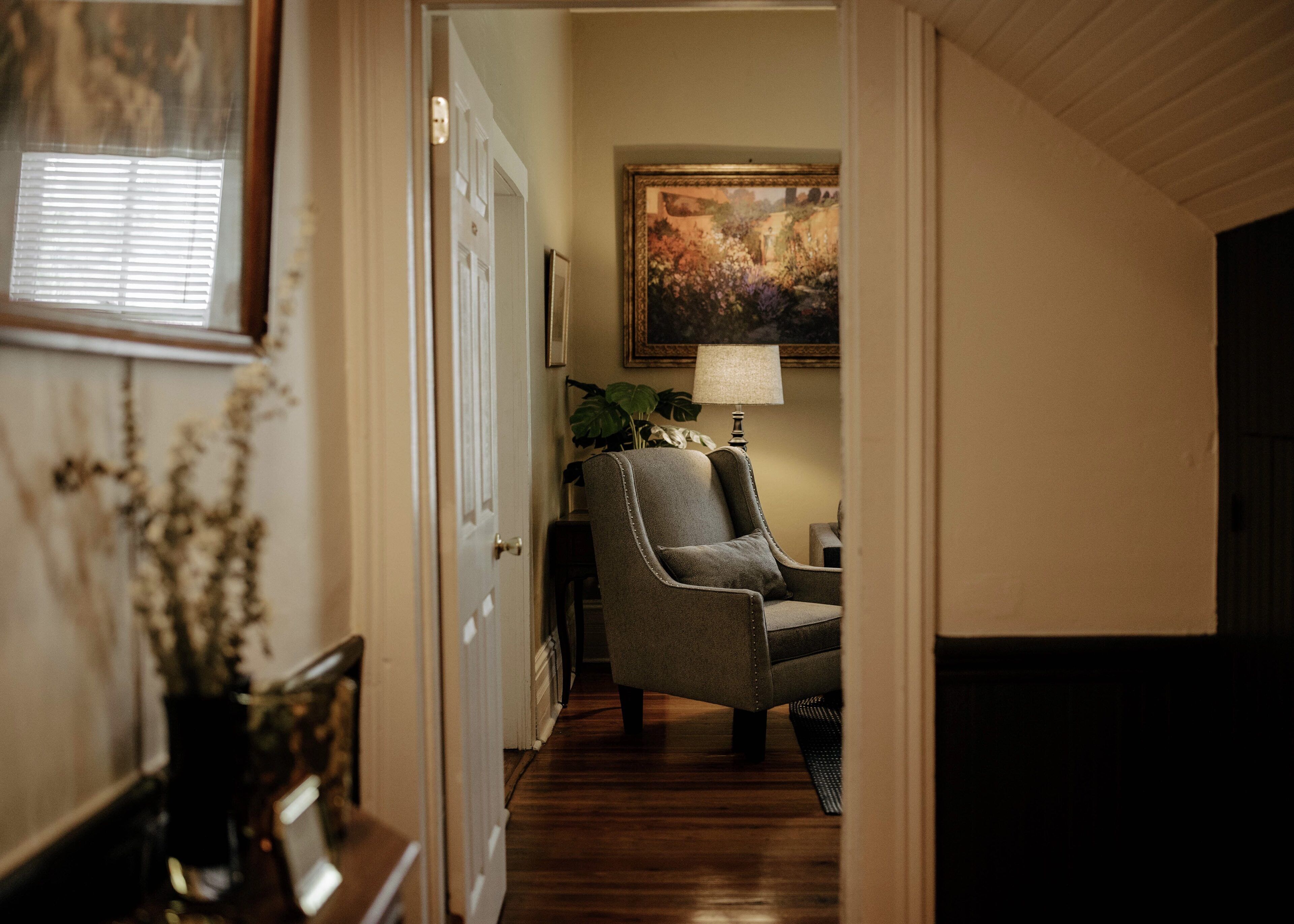 A hallway with a wooden floor, a gray armchair, a table with a lamp, and a painting on the wall.