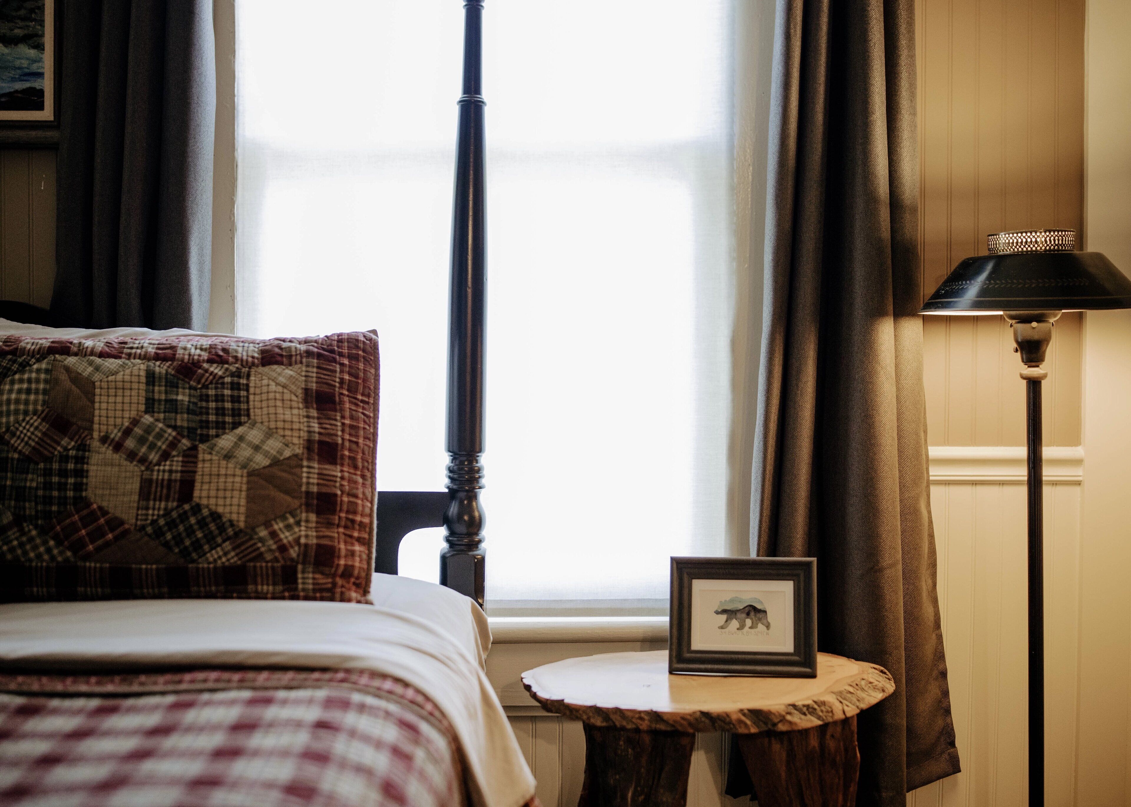 Una cama con una colcha escocesa, un cuadro enmarcado sobre una mesita de madera y una lámpara con pantalla estampada.