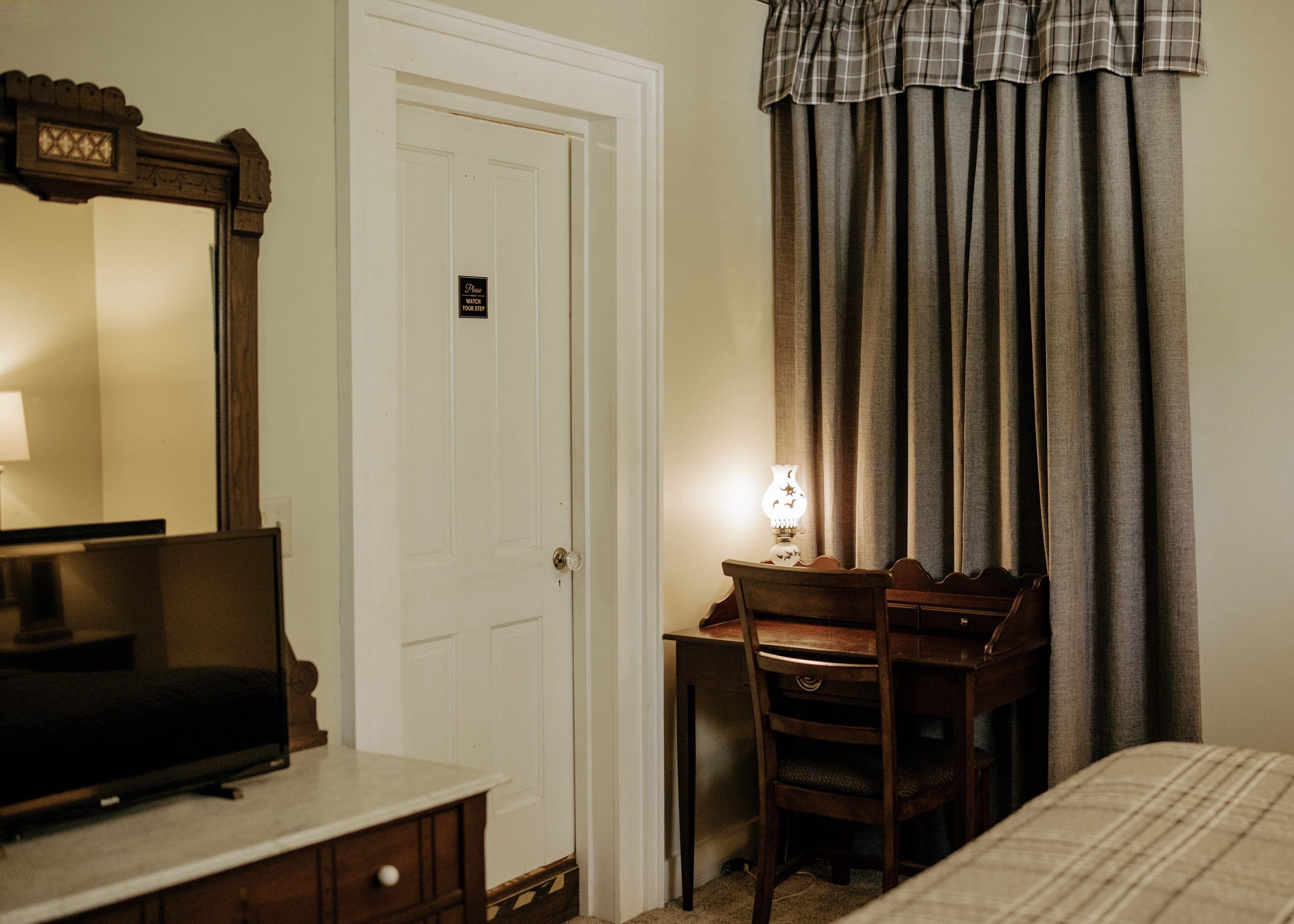 Une chambre d’hôtel avec un lit, un bureau et une chaise, un téléviseur posé sur une commode, une porte avec une pancarte et des rideaux.