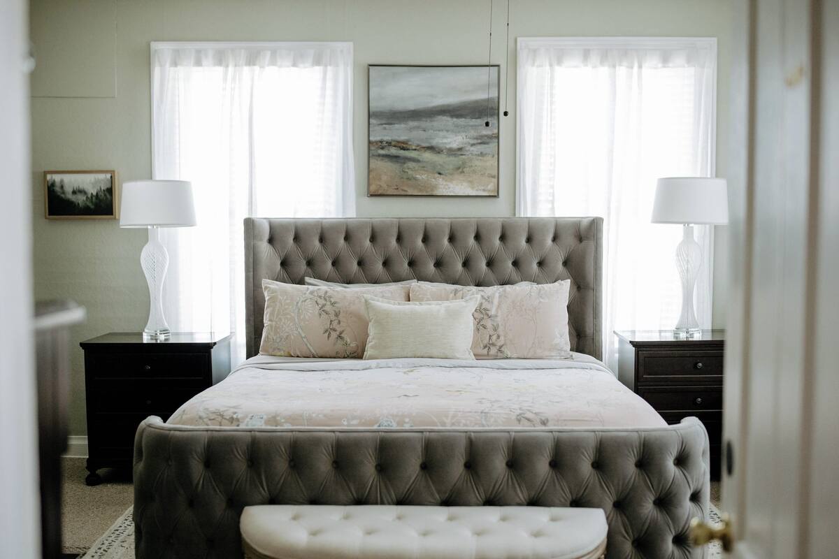 Quarto com cabeceira estofada, cama grande com roupa de cama floral, duas mesas de cabeceira com abajures e um banquinho ao pé da cama.