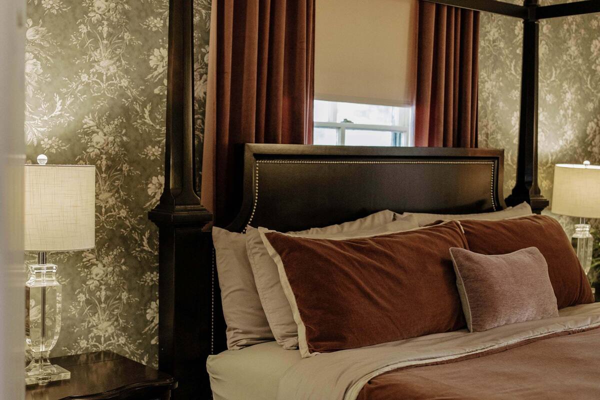 Uma cama com dossel, cabeceira escura, roupa de cama marrom e uma mesa de cabeceira com abajur.