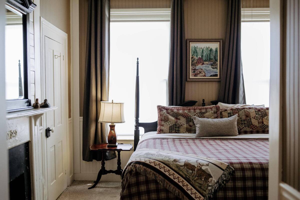 Quarto com cama, mesa de cabeceira, luminária, quadro e janela com cortinas.