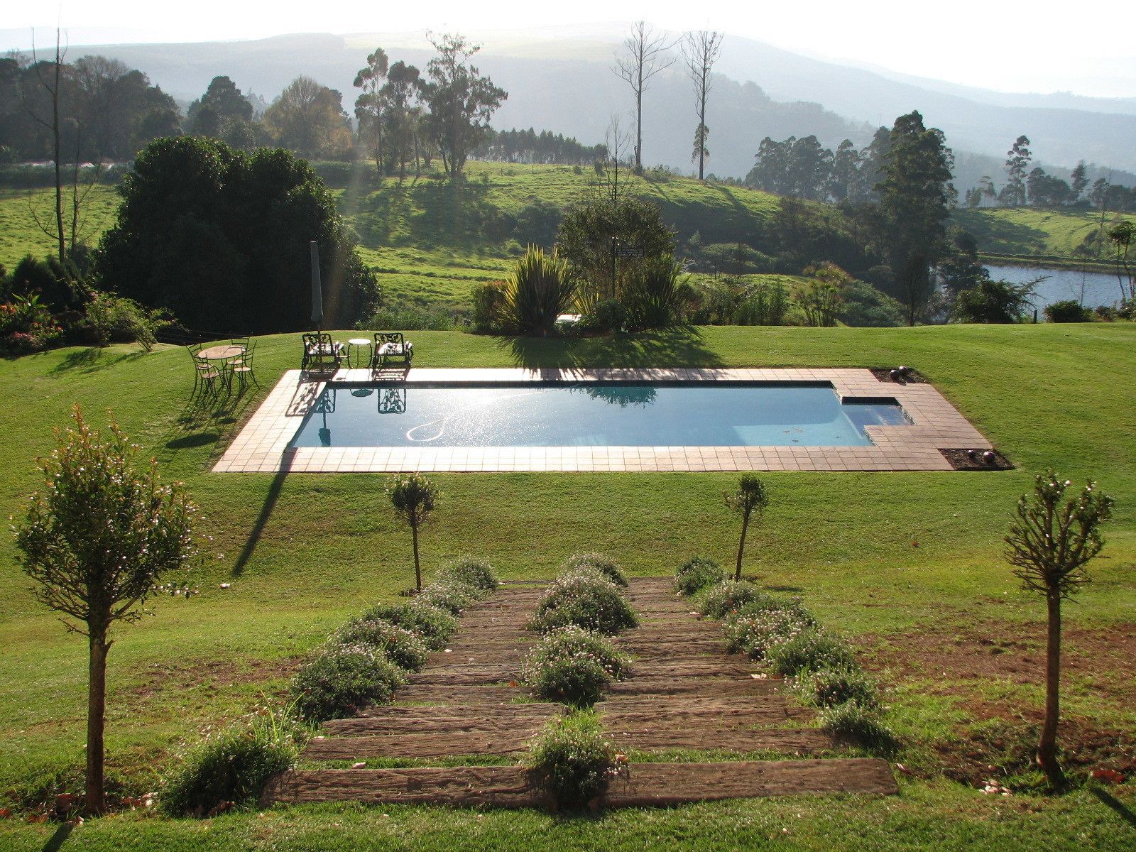 Une piscine rectangulaire, bordée d’une terrasse carrelée, entourée d’une pelouse bien entretenue et accessible par un chemin en bois.