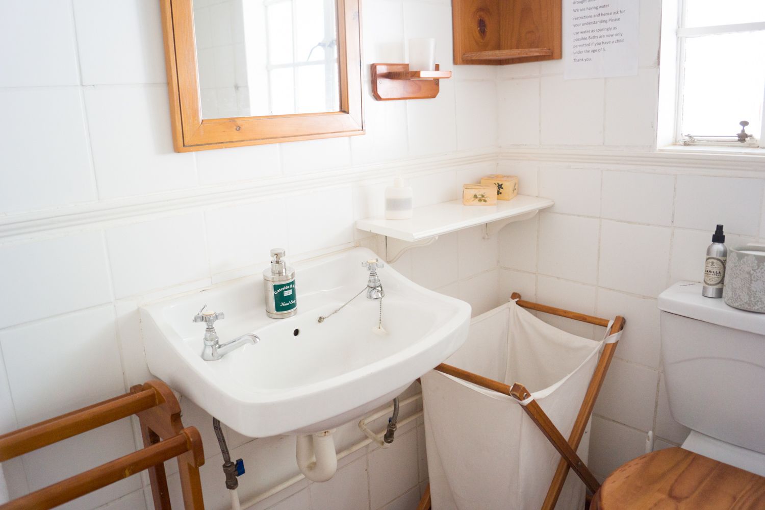 A bathroom with a white sink, two faucets, a mirror, a wooden shelf with toiletries, and a wooden chair.
