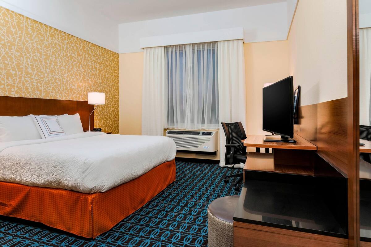 A hotel room with a bed, a desk with a TV, an air conditioner, and a patterned carpet.