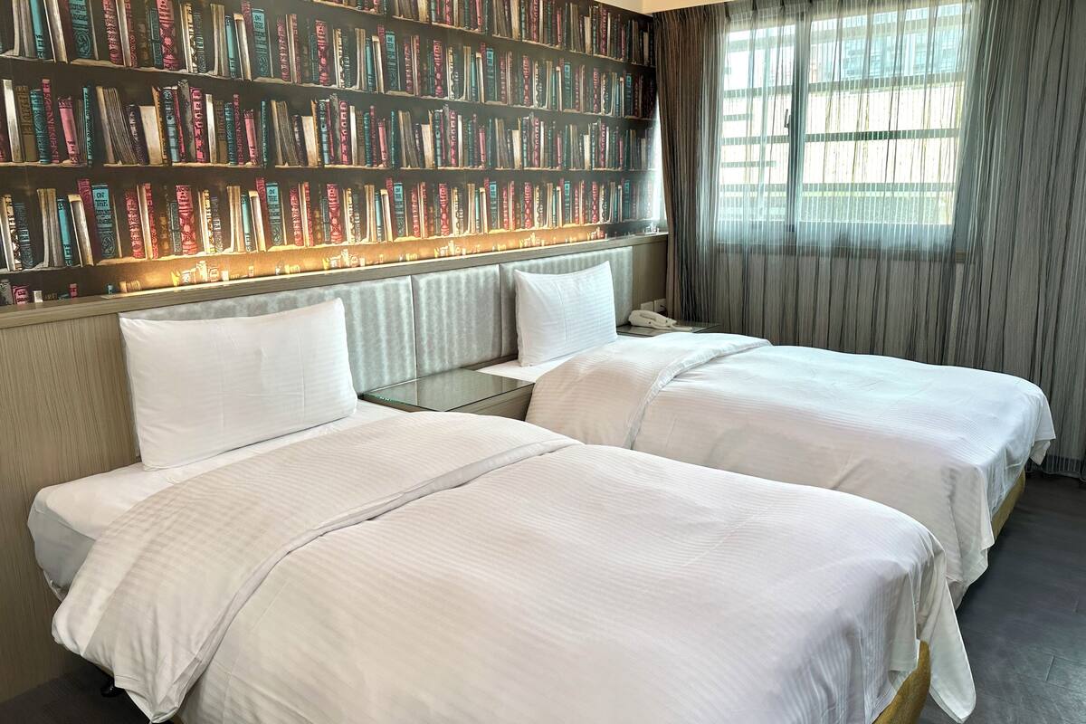 A hotel room with two beds, a bookshelf with books, a window with curtains, and a wooden headboard.