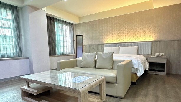 A modern hotel room with a sofa, a glass coffee table, a bed with white bedding, a nightstand, and a large window with curtains. - Beauty Trip Hotel (Taichung)