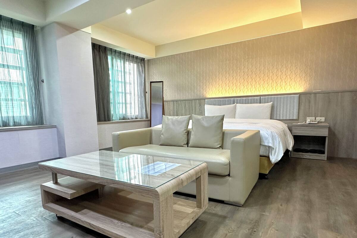 A modern hotel room with a sofa, a glass coffee table, a bed with white bedding, a nightstand, and a large window with curtains.