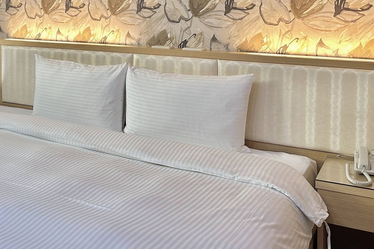 A hotel room with a bed, a bedside telephone, and a window with sheer curtains.