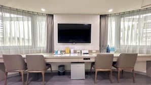 A hotel room with a large flat-screen TV mounted on the wall, a desk with four chairs, a small refrigerator, and a trash bin. - Beauty Trip Hotel (Taichung)