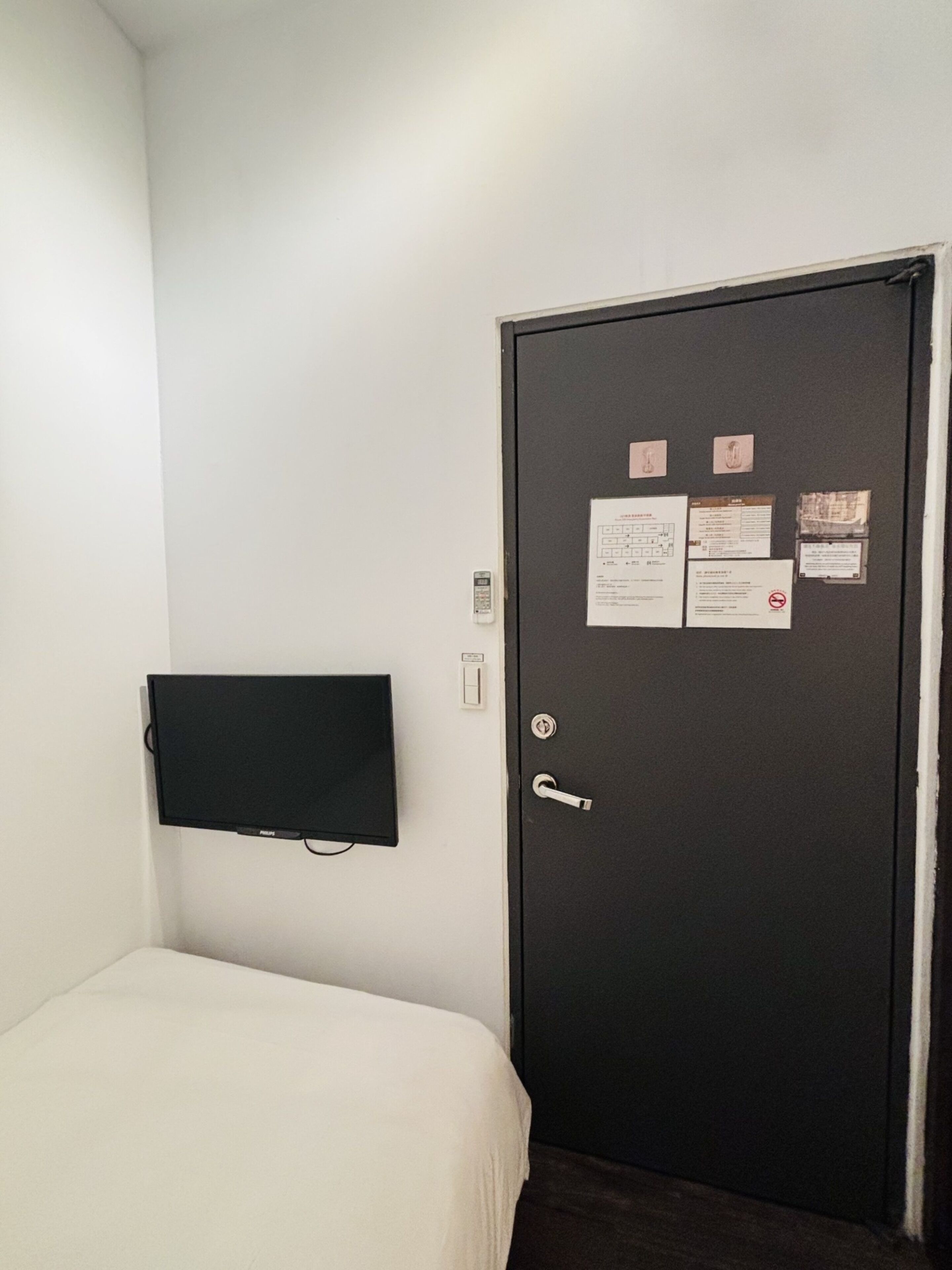 Ein Zimmer mit einem Bett, einem flachbildfernseher an der Wand und einer Tür mit mehreren Hinweisen.