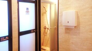 A hotel bathroom with a glass shower enclosure, a white towel dispenser, and a door with a frosted glass panel.
