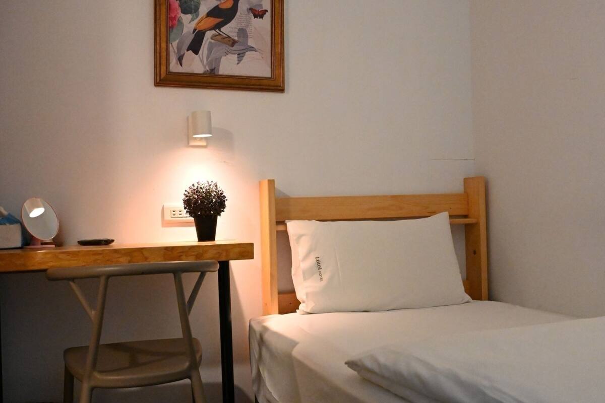 Une petite pièce, bien rangée, avec un lit, un bureau et une chaise. Un tableau encadré est accroché au mur, au-dessus du bureau.