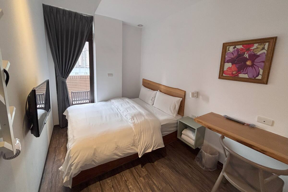 Une chambre d’hôtel comprenant un lit, un bureau, une chaise, une fenêtre avec des rideaux, un téléviseur fixé au mur et un tableau représentant des fleurs.