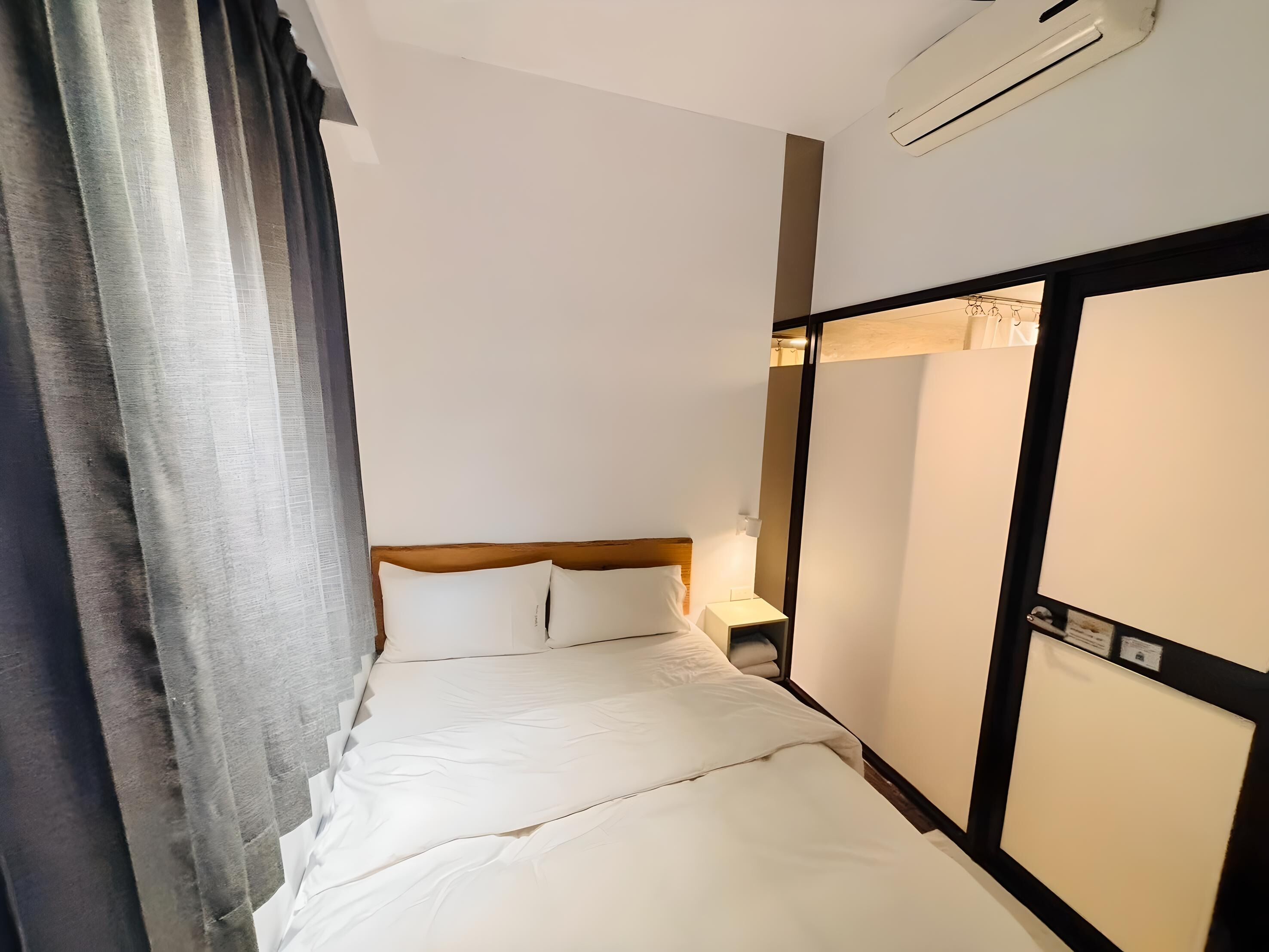 A hotel room with a bed, a nightstand, a wall-mounted air conditioner, and a window with curtains.