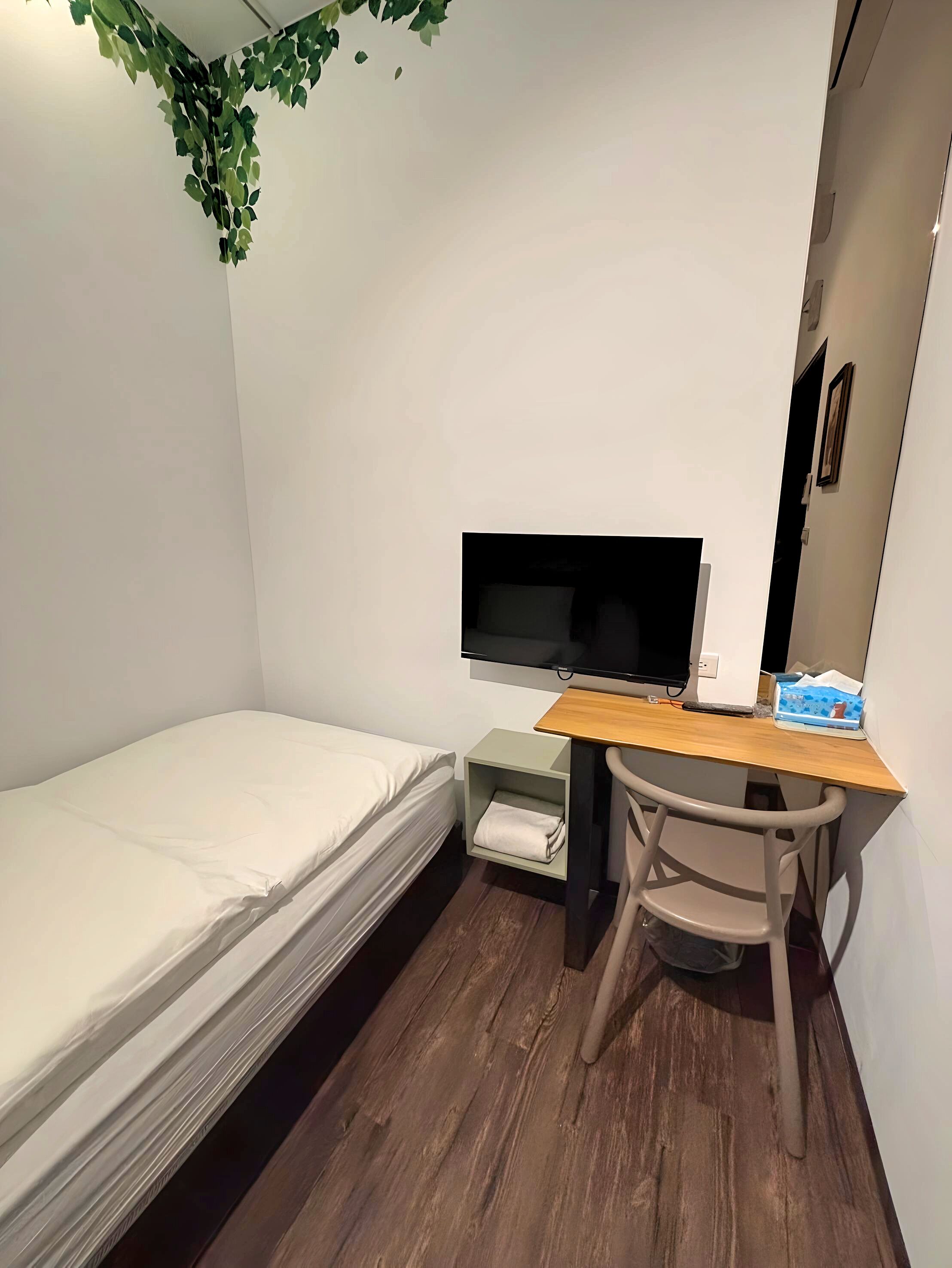 Une petite pièce, bien rangée, avec un lit simple, un bureau avec une chaise, un téléviseur à écran plat et une petite étagère.