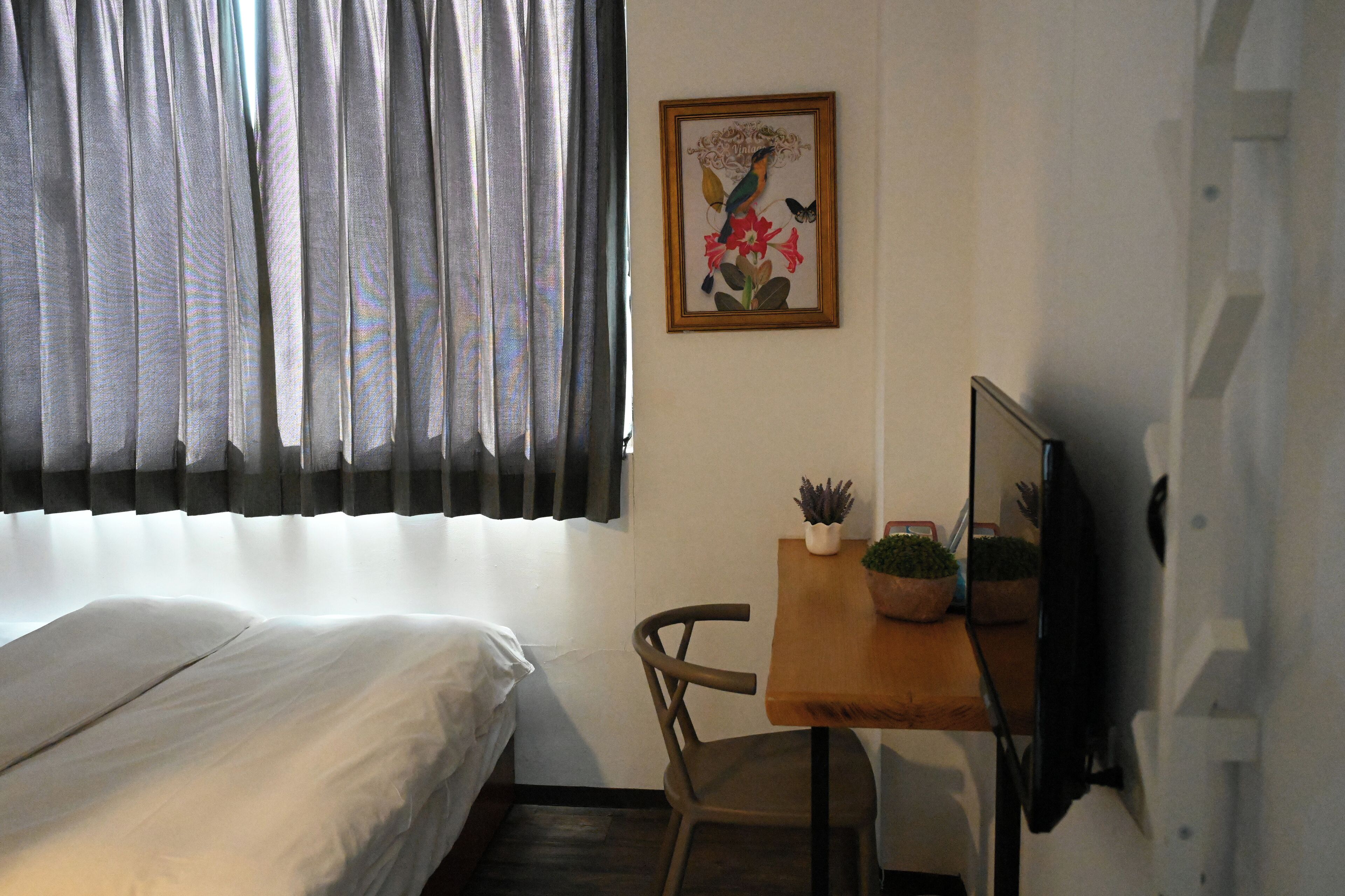 Una habitación con cama, un escritorio con planta en maceta, un cuadro en la pared y una ventana con cortinas.