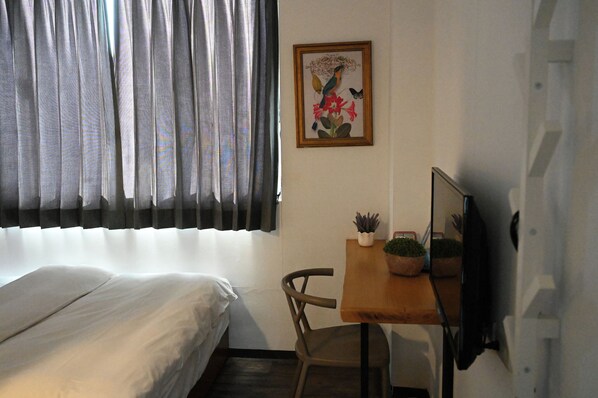 A room with a bed, a desk with a potted plant, a framed picture on the wall, and a window with curtains.