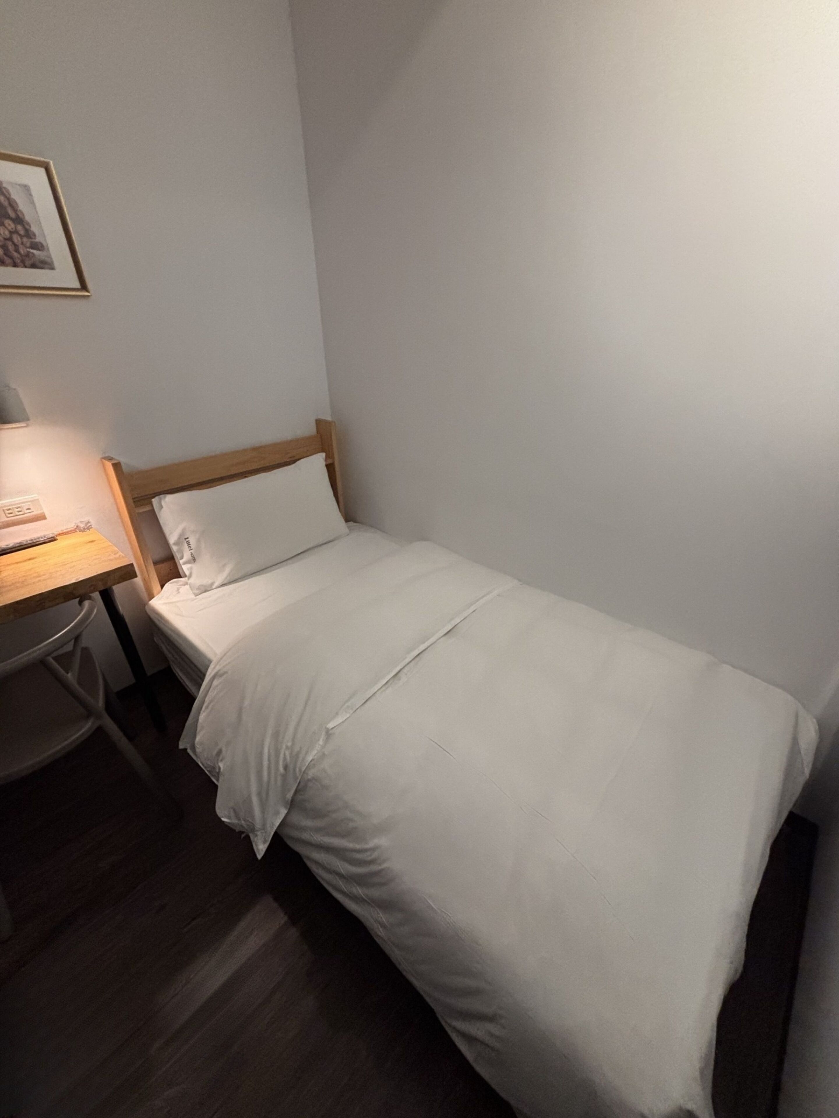A single bed with white bedding in a room with a wooden headboard. A desk with a lamp and a chair is to the left of the bed.