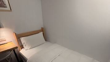 A single bed with white bedding in a room with a wooden headboard. A desk with a lamp and a chair is to the left of the bed.