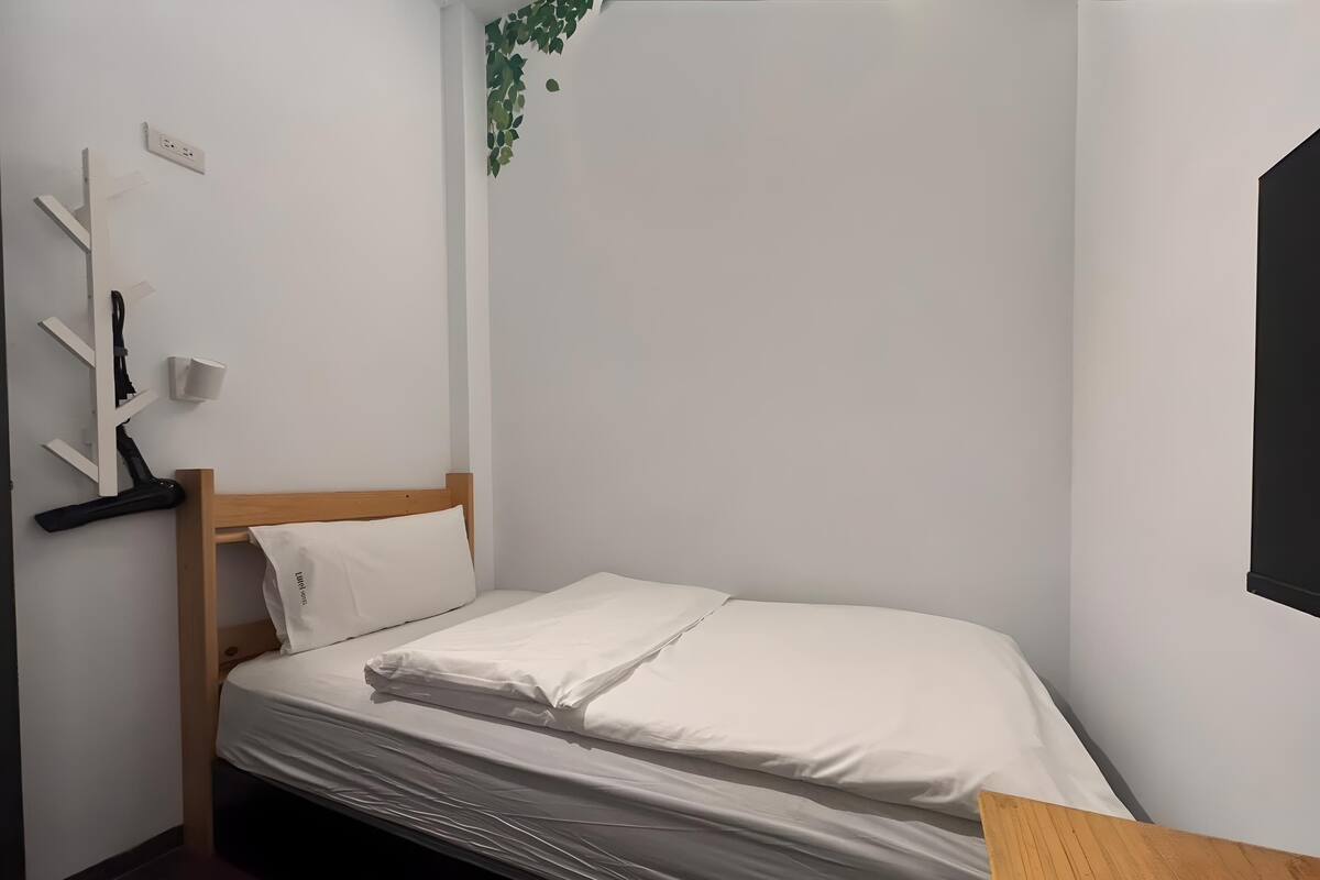 Un petit lit, bien fait, se trouve dans un coin de la pièce, contre un mur blanc. Il y a une prise de courant et une petite étagère avec une lampe.